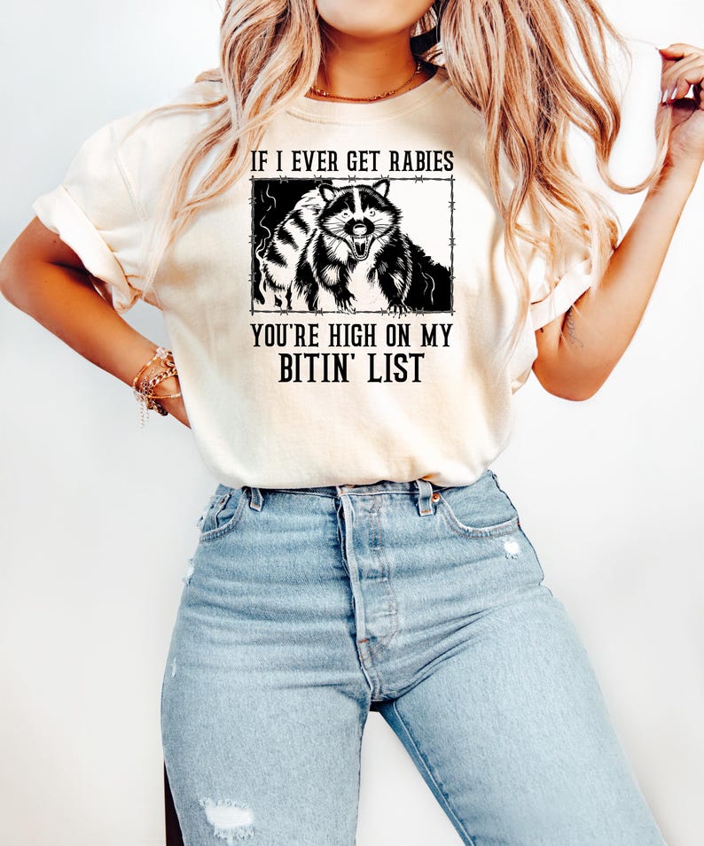 May include: Cream-colored t-shirt featuring a black and white raccoon graphic within a rectangular frame. The text reads, "IF I EVER GET RABIES YOU'RE HIGH ON MY BITIN' LIST."