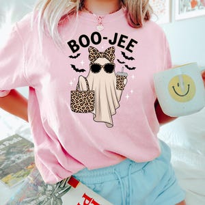 May include: Pink t-shirt with a graphic of a ghost wearing sunglasses and a leopard print bow, holding a leopard print tote bag and a coffee cup. The text "BOO-JEE" is above the ghost, with bats and sparkles. The person is also holding a mug with a smiley face.