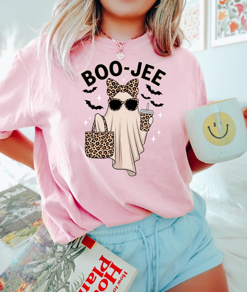 May include: Pink t-shirt with a graphic of a ghost wearing sunglasses and a leopard print bow, holding a leopard print tote bag and a coffee cup. The text "BOO-JEE" is above the ghost, with bats and sparkles. The person is also holding a mug with a smiley face.