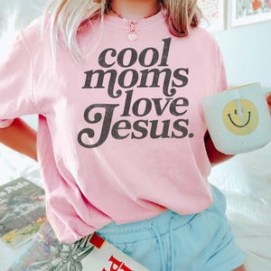 May include: A light pink sweatshirt with the text "cool moms love Jesus" in a black, bold font. The person is also holding a white mug with a yellow smiley face.