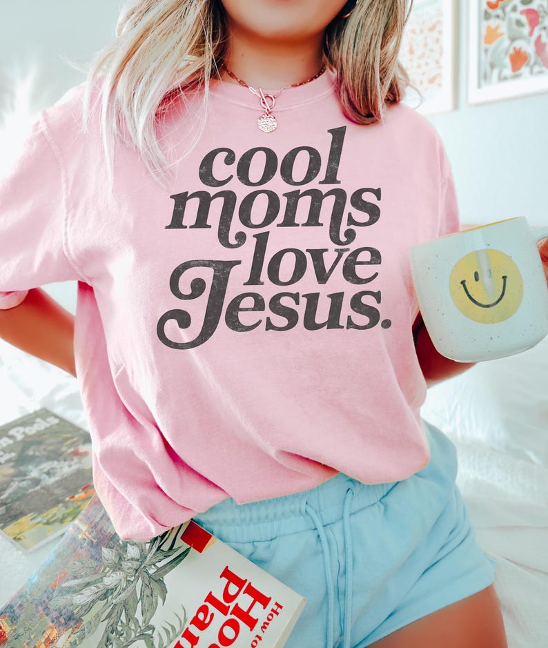 May include: A light pink sweatshirt with the text "cool moms love Jesus" in a black, bold font. The person is also holding a white mug with a yellow smiley face.