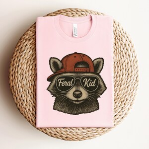May include: A light pink t-shirt featuring a raccoon wearing a brown baseball cap and sunglasses. The sunglasses have the words "Feral Kid" printed on them. The shirt is folded on a woven, circular surface.