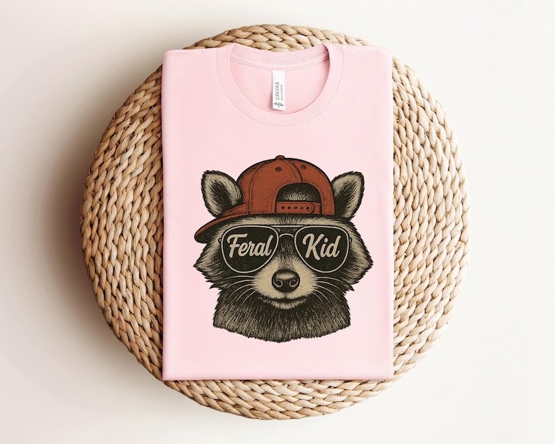 May include: A light pink t-shirt featuring a raccoon wearing a brown baseball cap and sunglasses. The sunglasses have the words "Feral Kid" printed on them. The shirt is folded on a woven, circular surface.