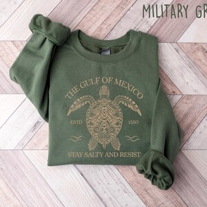 May include: A green sweatshirt with a gold graphic of a sea turtle with the text "The Gulf of Mexico" above it and "Stay Salty and Resist" below it. The text "Estd 1550" is also included on the graphic.