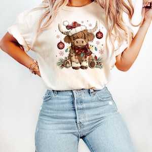 May include: Cream-colored t-shirt featuring a festive Highland cow illustration. The cow wears a Santa hat, a plaid scarf, and is adorned with Christmas lights and ornaments. The design is set against a backdrop of snowflakes and greenery.