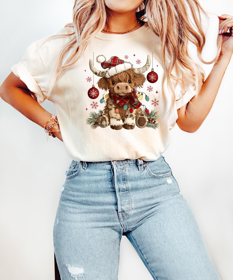 May include: Cream-colored t-shirt featuring a festive Highland cow illustration. The cow wears a Santa hat, a plaid scarf, and is adorned with Christmas lights and ornaments. The design is set against a backdrop of snowflakes and greenery.