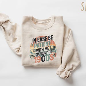 May include: A sand-colored sweatshirt with the text "PLEASE BE PATIENT WITH ME I'M FROM THE 1900S" in a retro design. The graphic includes images of a rotary phone, a television, and a radio.