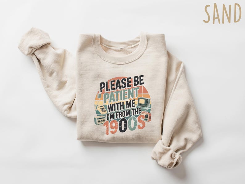 May include: A sand-colored sweatshirt with the text "PLEASE BE PATIENT WITH ME I'M FROM THE 1900S" in a retro design. The graphic includes images of a rotary phone, a television, and a radio.