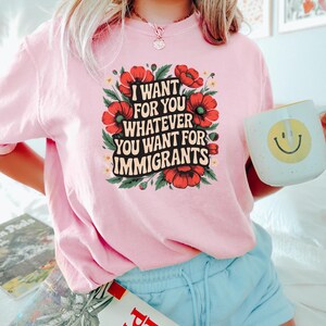 May include: A light pink t-shirt with the text "I WANT FOR YOU WHATEVER YOU WANT FOR IMMIGRANTS" surrounded by red flowers and green leaves. The person is holding a white mug with a yellow smiley face.