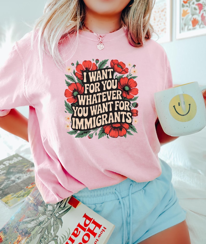 May include: A light pink t-shirt with the text "I WANT FOR YOU WHATEVER YOU WANT FOR IMMIGRANTS" surrounded by red flowers and green leaves. The person is holding a white mug with a yellow smiley face.