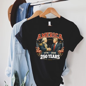 Puede incluir: Camiseta negra con un gr&aacute;fico de dos hombres d&aacute;ndose la mano, con la palabra "AMERICA" arriba y "1776-2026 250 YEARS" abajo. El dise&ntilde;o incluye banderas americanas y estrellas. La camiseta est&aacute; colgada en una percha de madera.