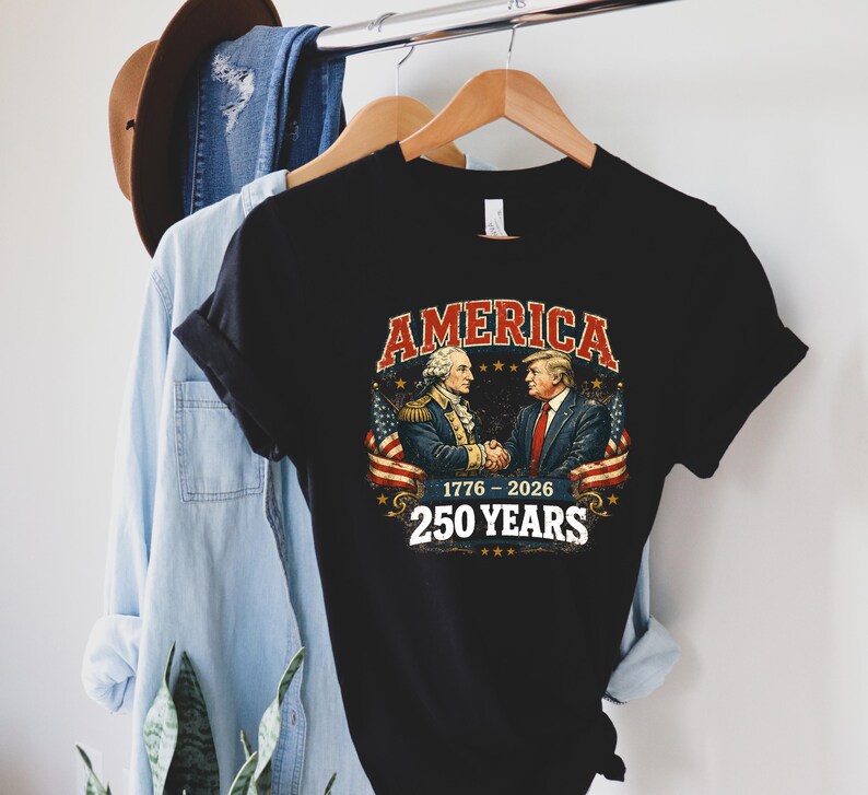 Puede incluir: Camiseta negra con un gr&aacute;fico de dos hombres d&aacute;ndose la mano, con la palabra "AMERICA" arriba y "1776-2026 250 YEARS" abajo. El dise&ntilde;o incluye banderas americanas y estrellas. La camiseta est&aacute; colgada en una percha de madera.