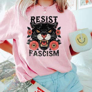 May include: A light pink t-shirt with the words "RESIST FASCISM" above a black panther graphic with red and orange flowers. The person is holding a white mug with a yellow smiley face.
