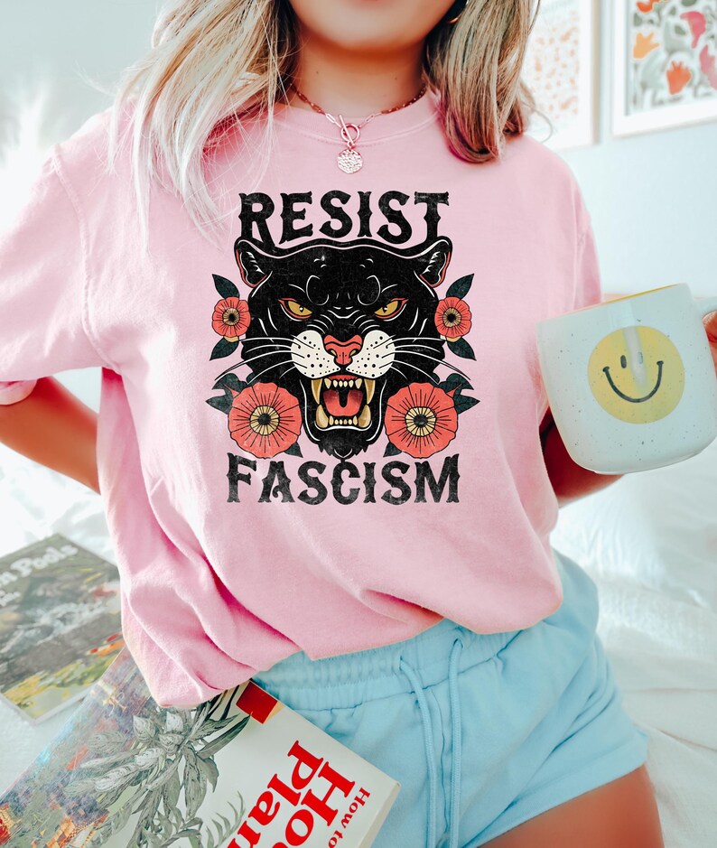 May include: A light pink t-shirt with the words "RESIST FASCISM" above a black panther graphic with red and orange flowers. The person is holding a white mug with a yellow smiley face.