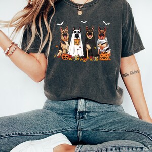May include: Dark gray t-shirt featuring a Halloween-themed graphic. The design includes four German Shepherd dogs dressed in costumes, with pumpkins and bats. The shirt is a comfortable, casual style.
