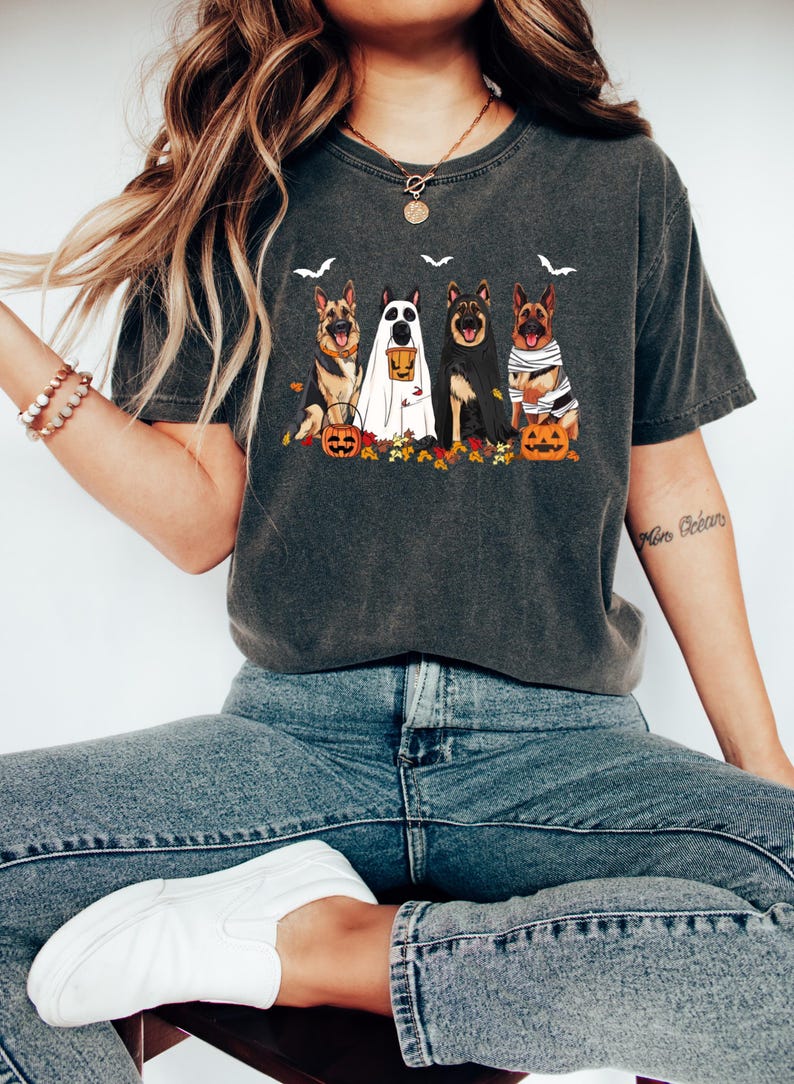 May include: Dark gray t-shirt featuring a Halloween-themed graphic. The design includes four German Shepherd dogs dressed in costumes, with pumpkins and bats. The shirt is a comfortable, casual style.