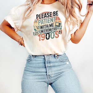 May include: Cream-colored t-shirt with the text "PLEASE BE PATIENT WITH ME I'M FROM THE 1900S" in a retro design. The graphic includes vintage electronics like a TV, radio, and telephone. The shirt is paired with light blue denim jeans.