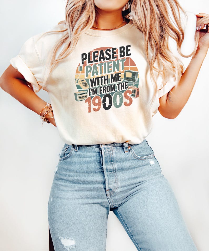 May include: Cream-colored t-shirt with the text "PLEASE BE PATIENT WITH ME I'M FROM THE 1900S" in a retro design. The graphic includes vintage electronics like a TV, radio, and telephone. The shirt is paired with light blue denim jeans.