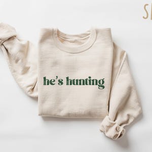 May include: A sand-colored sweatshirt with the text "he's hunting" in a distressed green font. The long sleeves are rolled up. The sweatshirt is made of a soft material.