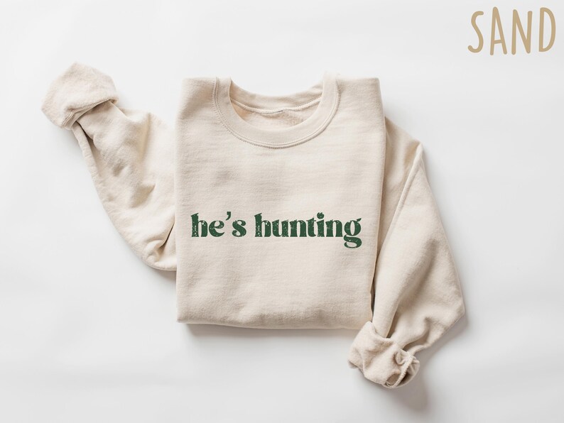 May include: A sand-colored sweatshirt with the text "he's hunting" in a distressed green font. The long sleeves are rolled up. The sweatshirt is made of a soft material.