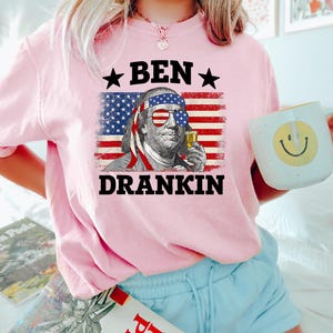 May include: Pink t-shirt featuring a graphic of Benjamin Franklin wearing sunglasses and holding a drink, set against an American flag backdrop. The text "BEN DRANKIN" is above and below the image. The person is also holding a white mug with a yellow smiley face.