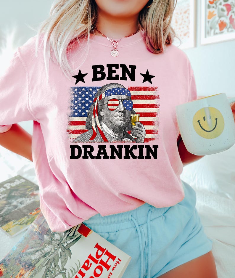 May include: Pink t-shirt featuring a graphic of Benjamin Franklin wearing sunglasses and holding a drink, set against an American flag backdrop. The text "BEN DRANKIN" is above and below the image. The person is also holding a white mug with a yellow smiley face.