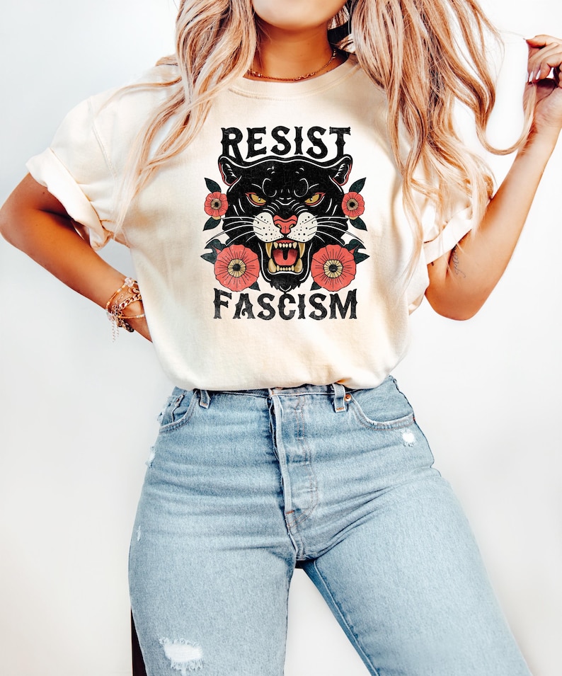 May include: Cream-colored t-shirt featuring a black panther graphic with the words "RESIST FASCISM" in bold letters. The panther is surrounded by red flowers. The shirt is paired with light blue jeans.