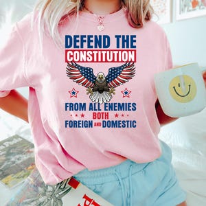 May include: Pink t-shirt with the text "DEFEND THE CONSTITUTION" above an American flag eagle graphic. The text "FROM ALL ENEMIES BOTH FOREIGN AND DOMESTIC" is below the eagle. The person is holding a white mug with a yellow smiley face.