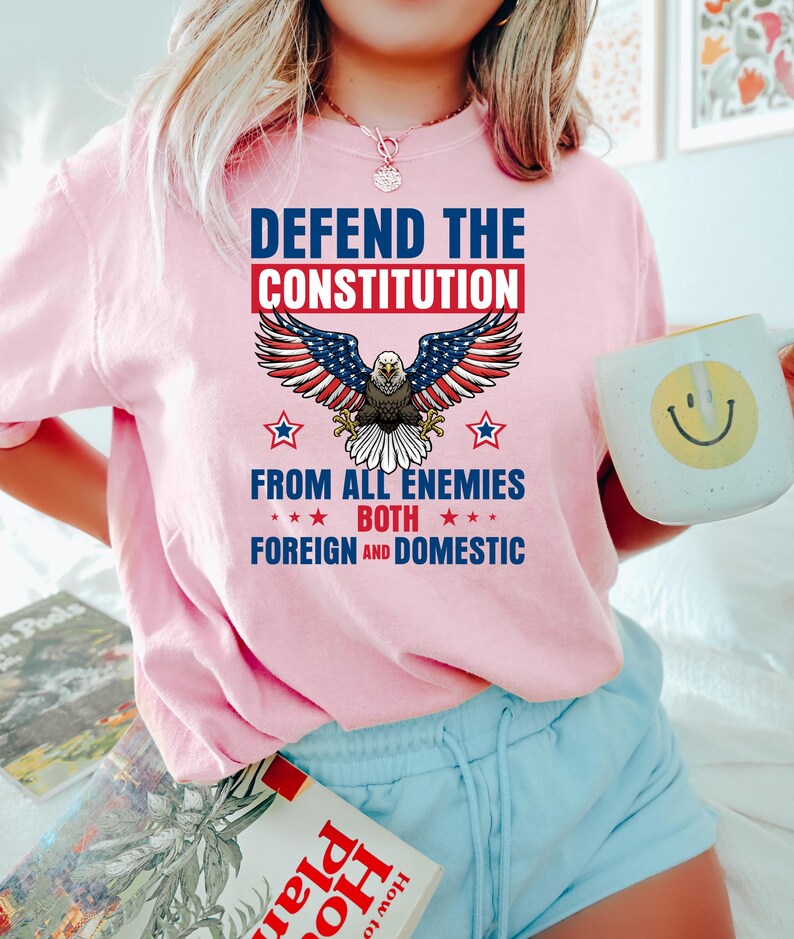 May include: Pink t-shirt with the text "DEFEND THE CONSTITUTION" above an American flag eagle graphic. The text "FROM ALL ENEMIES BOTH FOREIGN AND DOMESTIC" is below the eagle. The person is holding a white mug with a yellow smiley face.