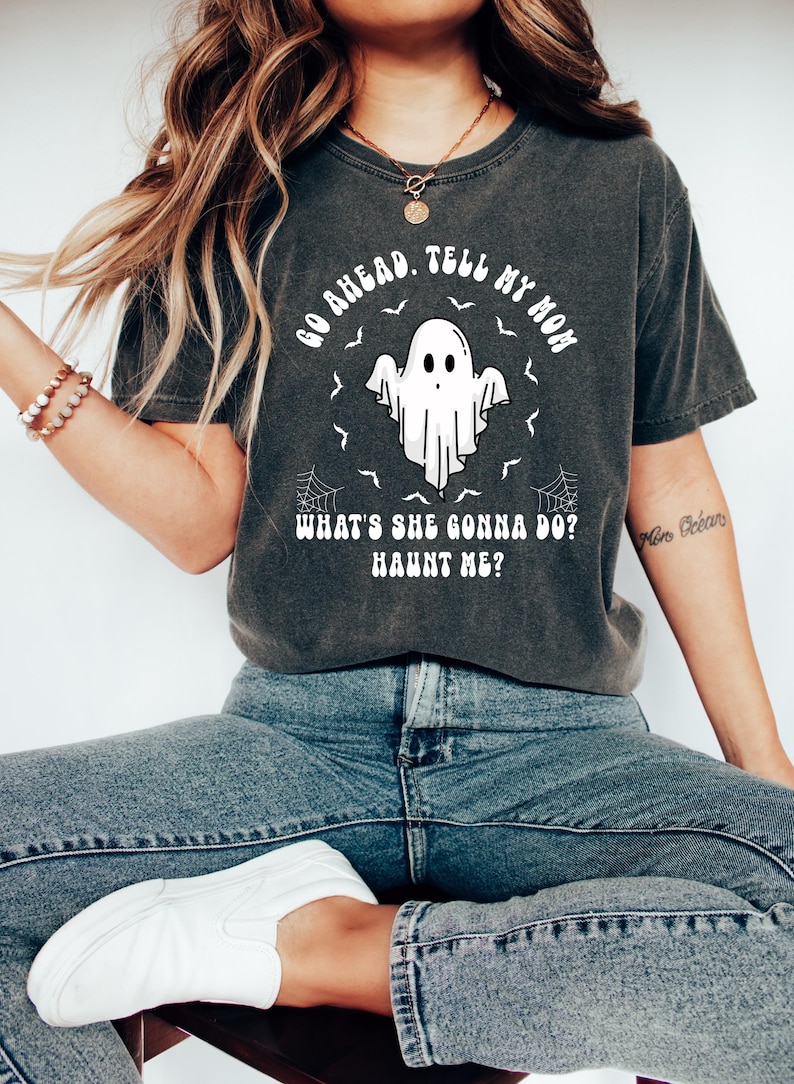 May include: Dark grey t-shirt featuring a white ghost graphic and the text "GO AHEAD. TELL MY MOM WHAT'S SHE GONNA DO? HAUNT ME?". The shirt is worn with blue jeans and white shoes. A Halloween-themed top.