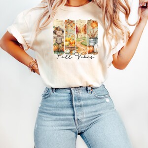 May include: Cream-colored t-shirt featuring a fall-themed graphic with the text "Fall Vibes." The graphic includes images of a scarecrow, pumpkins, and autumn foliage. The shirt is paired with light blue jeans.
