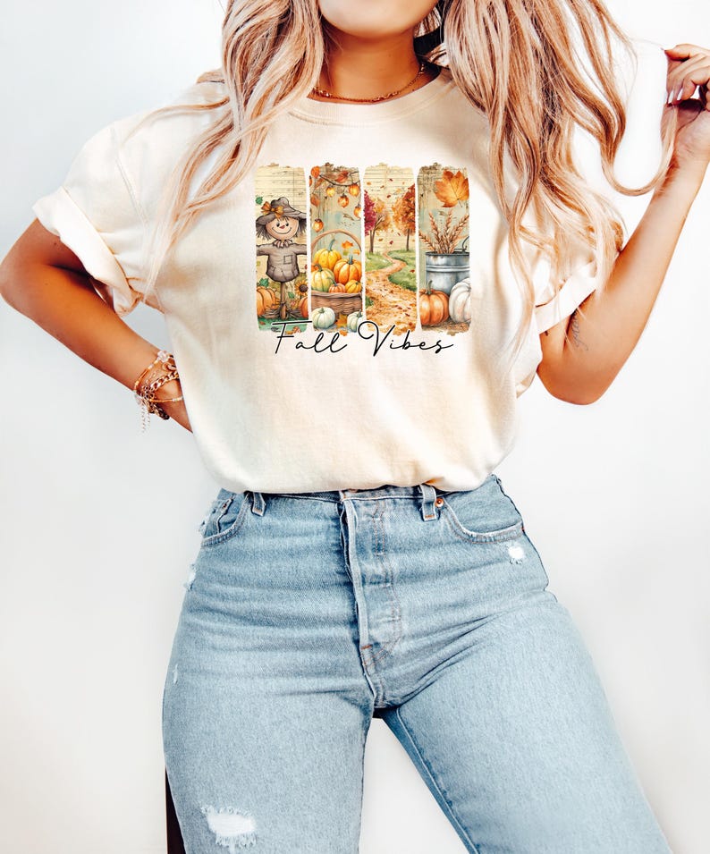 May include: Cream-colored t-shirt featuring a fall-themed graphic with the text "Fall Vibes." The graphic includes images of a scarecrow, pumpkins, and autumn foliage. The shirt is paired with light blue jeans.