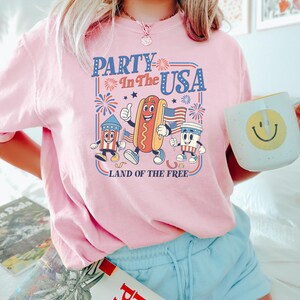 May include: Pink t-shirt with a retro graphic that reads "Party in the USA" featuring cartoon hot dog, firecracker, and coffee cup characters. The shirt also has the text "Land of the Free". The person is holding a mug with a smiley face.