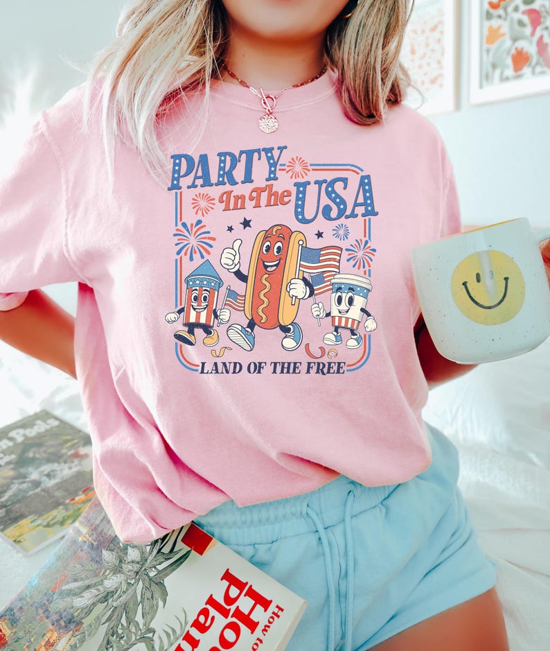 May include: Pink t-shirt with a retro graphic that reads "Party in the USA" featuring cartoon hot dog, firecracker, and coffee cup characters. The shirt also has the text "Land of the Free". The person is holding a mug with a smiley face.