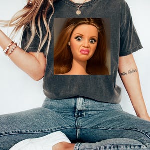 May include: Dark gray t-shirt featuring a doll's face with a surprised expression. The doll has long brown hair, large eyes, and a puckered mouth. The shirt is paired with blue jeans and white sneakers. A gold necklace and bracelet are also visible.