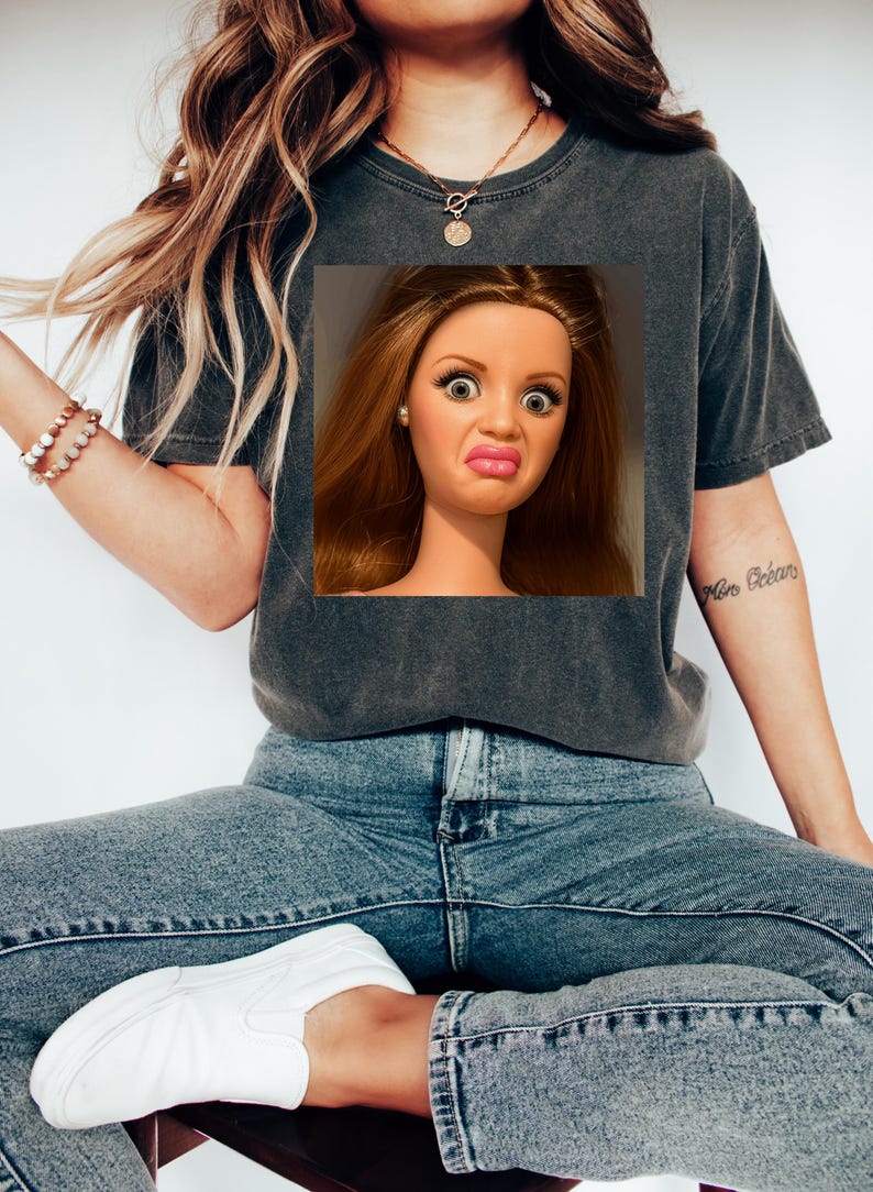 May include: Dark gray t-shirt featuring a doll's face with a surprised expression. The doll has long brown hair, large eyes, and a puckered mouth. The shirt is paired with blue jeans and white sneakers. A gold necklace and bracelet are also visible.
