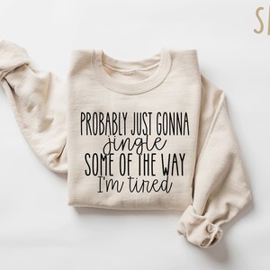 Op de afbeelding: Een zandkleurige sweatshirt met de zwarte tekst "PROBABLY JUST GONNA jingle SOME OF THE WAY I'M tired". De mouwen zijn opgevouwen en de tekst staat in het midden van de voorkant. Het woord "SAND" staat rechtsboven.