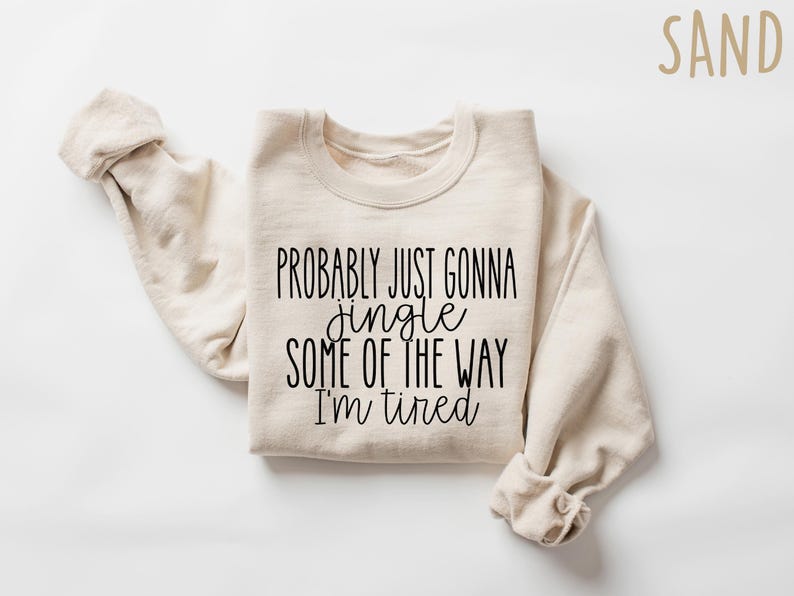Op de afbeelding: Een zandkleurige sweatshirt met de zwarte tekst "PROBABLY JUST GONNA jingle SOME OF THE WAY I'M tired". De mouwen zijn opgevouwen en de tekst staat in het midden van de voorkant. Het woord "SAND" staat rechtsboven.