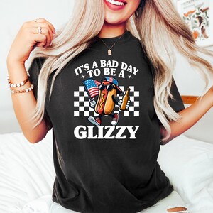 May include: Black t-shirt with a white graphic of a hot dog holding an American flag and a beer. The text on the shirt reads "It's a bad day to be a Glizzy".