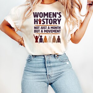 Puede incluir: Camiseta color crema con el texto "WOMEN'S HISTORY" y "NOT JUST A MONTH BUT A MOVEMENT" en morado oscuro. El dise&ntilde;o incluye un pu&ntilde;o en alto y siluetas de mujeres. La camiseta se combina con vaqueros azules.