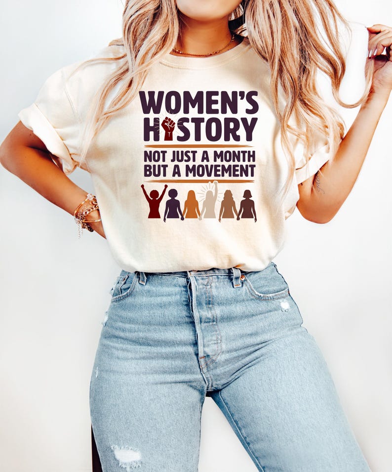 Puede incluir: Camiseta color crema con el texto "WOMEN'S HISTORY" y "NOT JUST A MONTH BUT A MOVEMENT" en morado oscuro. El dise&ntilde;o incluye un pu&ntilde;o en alto y siluetas de mujeres. La camiseta se combina con vaqueros azules.
