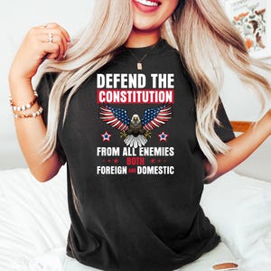 May include: Black t-shirt with the text "DEFEND THE CONSTITUTION" above an eagle graphic with red, white, and blue wings. The text "FROM ALL ENEMIES BOTH FOREIGN AND DOMESTIC" is below the eagle. The shirt has a crew neck.