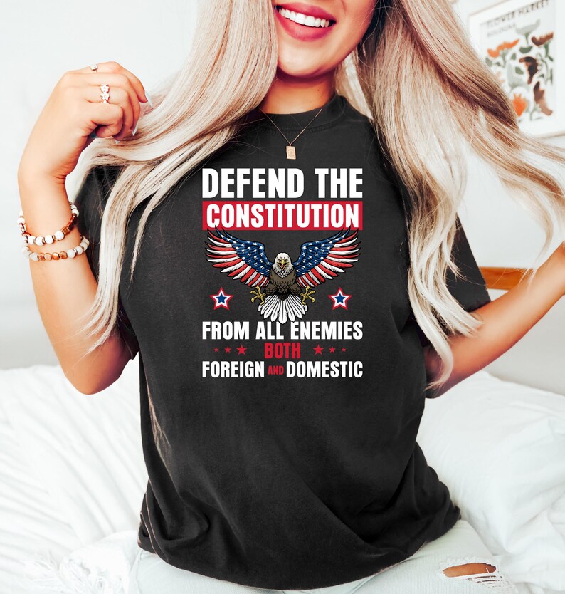 May include: Black t-shirt with the text "DEFEND THE CONSTITUTION" above an eagle graphic with red, white, and blue wings. The text "FROM ALL ENEMIES BOTH FOREIGN AND DOMESTIC" is below the eagle. The shirt has a crew neck.