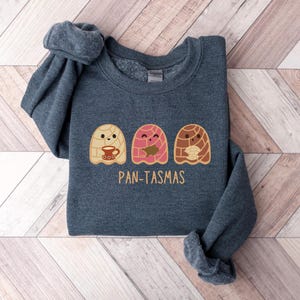 May include: A dark gray sweatshirt with a graphic of three cartoon pan dulce ghosts holding cups of coffee. The text "PAN-TASMAS" is printed below the ghosts.