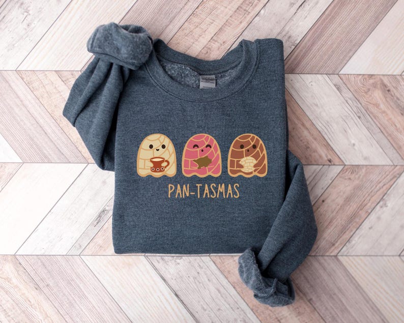 May include: A dark gray sweatshirt with a graphic of three cartoon pan dulce ghosts holding cups of coffee. The text "PAN-TASMAS" is printed below the ghosts.