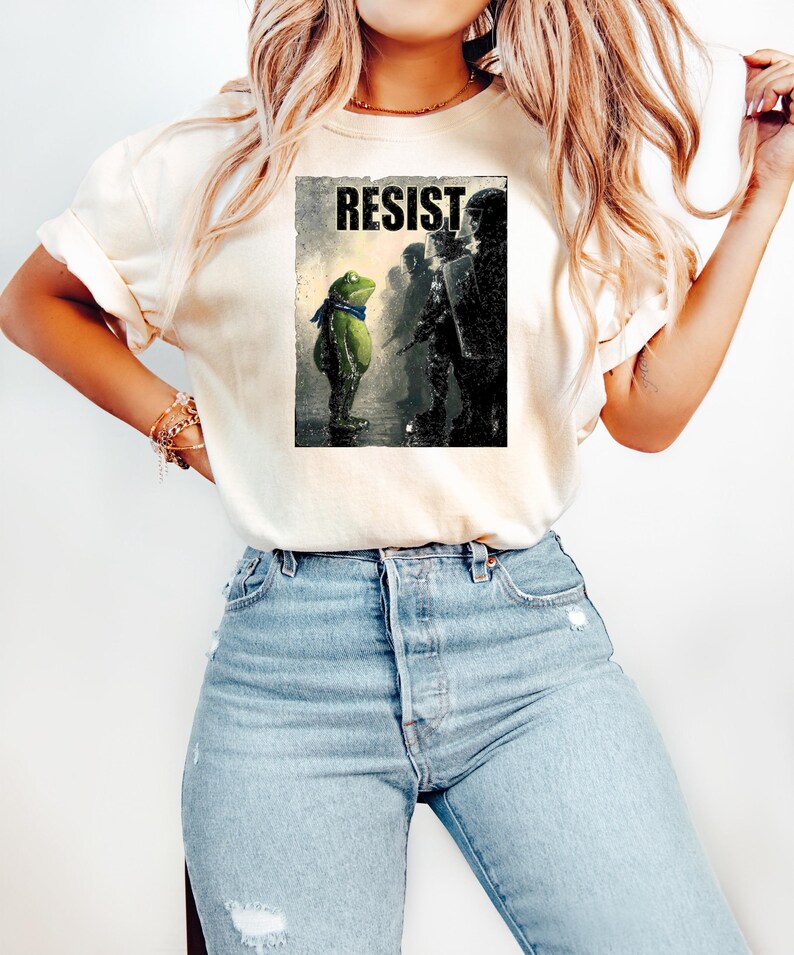 May include: Cream-colored t-shirt featuring a graphic of a frog wearing a blue scarf, facing a line of riot police. The word "RESIST" is printed above the frog. The shirt is paired with light blue jeans.