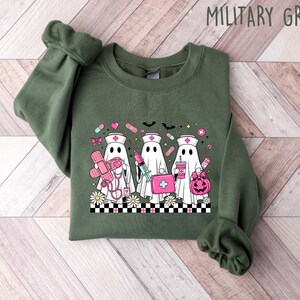 May include: Military green sweatshirt featuring a graphic of three ghost nurses with medical supplies and a pumpkin. The design includes a checkerboard pattern and daisies at the bottom. The text "MILITARY GREEN" is at the top.