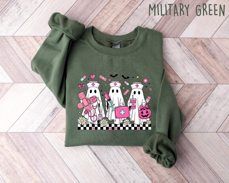 May include: Military green sweatshirt featuring a graphic of three ghost nurses with medical supplies and a pumpkin. The design includes a checkerboard pattern and daisies at the bottom. The text "MILITARY GREEN" is at the top.