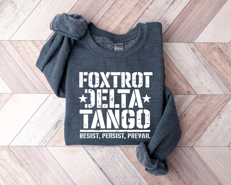 May include: A dark gray sweatshirt with the words "Foxtrot Delta Tango" in white block letters, with the words "Resist, Persist, Prevail" underneath. The sweatshirt is folded on a light brown wooden surface.