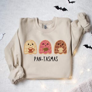 May include: A beige sweatshirt with a graphic of three cute ghosts made of pan dulce, a traditional Mexican sweet bread. The text "PAN-TASMAS" is printed below the ghosts.